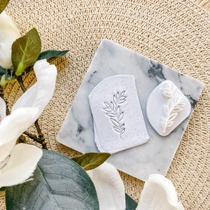 May include: Two white ceramic stamps with a leafy design on a white marble surface. The stamps are on a woven straw mat.