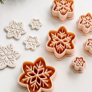 May include: A selection of snowflake-shaped cookie cutters in different sizes. The cutters are made of light pink plastic and are displayed on a white surface. The snowflakes feature intricate designs, ideal for festive baking.