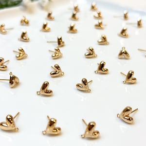May include: A selection of small, gold-coloured heart-shaped stud earrings displayed on a white surface. Each earring features a post for insertion and a small loop at the heart's base. The earrings are evenly spaced, creating a pattern.