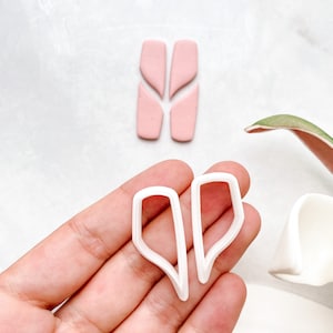 Peut inclure: Deux découpoirs en plastique blanc pour pâte polymère de forme unique sont tenus. Au-dessus, quatre morceaux d'argile rose sont disposés pour ressembler à la forme des découpoirs. Le fond est de couleur claire et neutre.