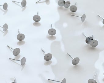 May include: A collection of silver-colored earring posts with flat, round pads and thin posts, scattered across a glossy white surface. The earring components are designed for jewelry making, with a simple, minimalist aesthetic.