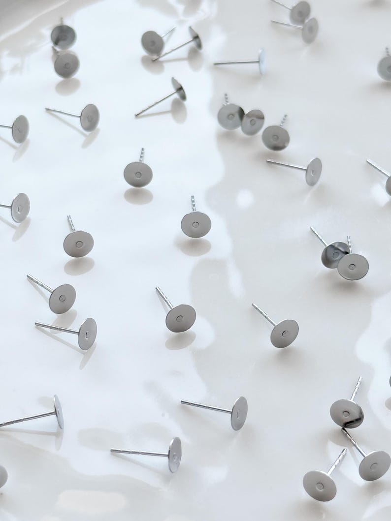 May include: Silver earring posts with flat, circular pads and slender stems are arranged on a shiny white surface. These components are for crafting earrings, featuring a minimalist design. The posts are made of metal.