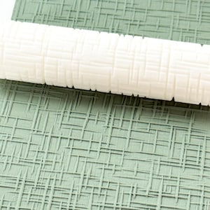 May include: A white textured rolling pin rests on a green surface with a grid pattern. The rolling pin has a similar grid design. The image showcases a tool for creating textured surfaces, likely for baking or crafting.