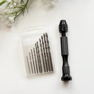 May include: A set of drill bits in a clear plastic case with a black drill bit holder. The drill bits are various sizes and are arranged in order of size. The drill bit holder has a black handle and a silver chuck.