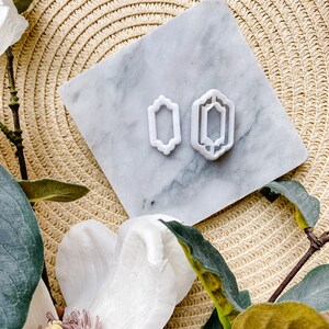 May include: Two white plastic cookie cutters in the shape of a rectangle with decorative edges. The cutters are on a white marble surface.