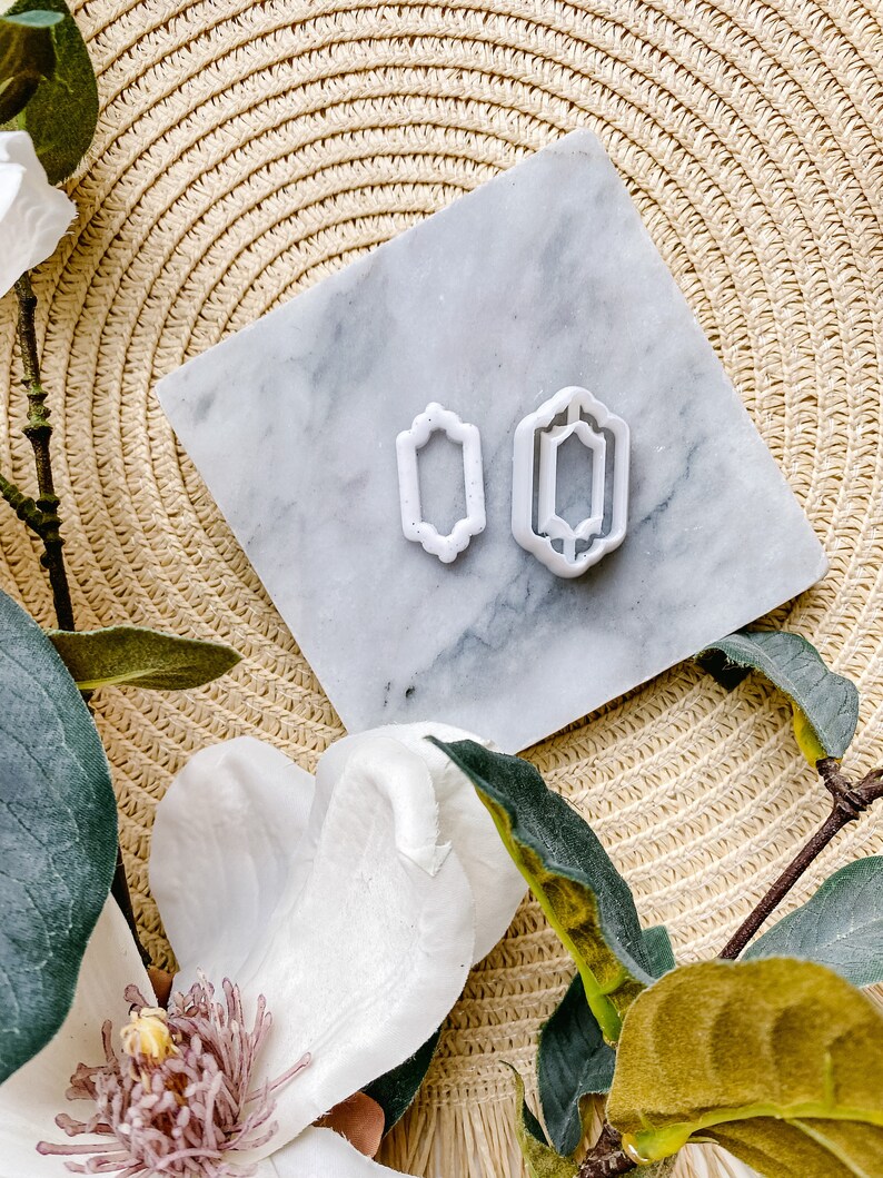 May include: Two white plastic cookie cutters in the shape of a rectangle with decorative edges. The cutters are on a white marble surface.