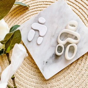 Könnte beinhalten: Zwei Sets weißer Polymer-Ton-Ohrringkomponenten auf einer Marmorplatte. Ein Set ist montiert, das andere ist unmontiert. Die Komponenten sind organische Formen mit gesprenkelten Details. Der Hintergrund ist eine gewebte Strohmatte.