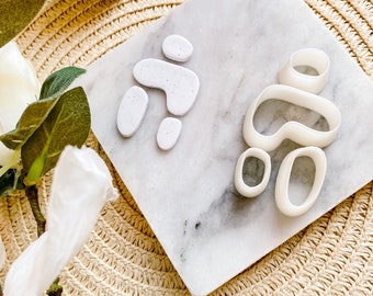 May include: Two sets of white polymer clay earring components on a marble tile. One set is assembled, the other is unassembled. The components are organic shapes with speckled details. The background is a woven straw mat.