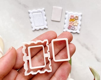 Può includere: Due tagliabiscotti in plastica bianca a forma di cornici. La cornice più grande ha un bordo ondulato e la cornice più piccola è liscia. I tagliabiscotti sono tenuti in una mano.