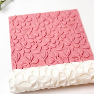 Könnte beinhalten: Eine rosa Prägeplatte mit einem floralen Hibiskusmuster und Blättern. Darunter befindet sich eine weiße Teigrolle mit passendem Blumendesign. Das Bild ist vor einem weißen Hintergrund aufgenommen.