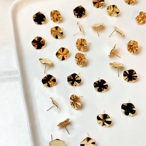 May include: A collection of 25 gold-toned metal earring studs with a scalloped edge design. The earrings are displayed on a white plate.