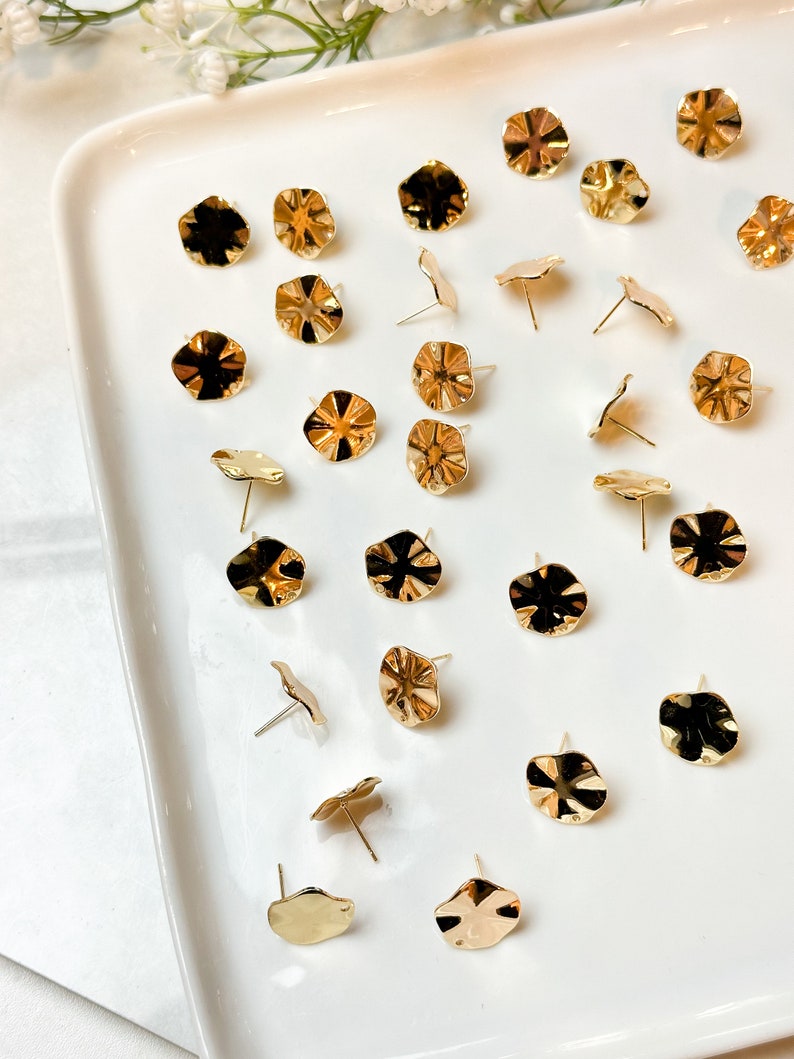 May include: A collection of 25 gold-toned metal earring studs with a scalloped edge design. The earrings are displayed on a white plate.