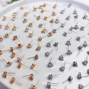 May include: A collection of gold-toned and silver-toned stud earrings, each featuring a single clear round gemstone set in a four-prong metal basket. Each earring includes a small loop at the base of the setting for attaching additional charms.