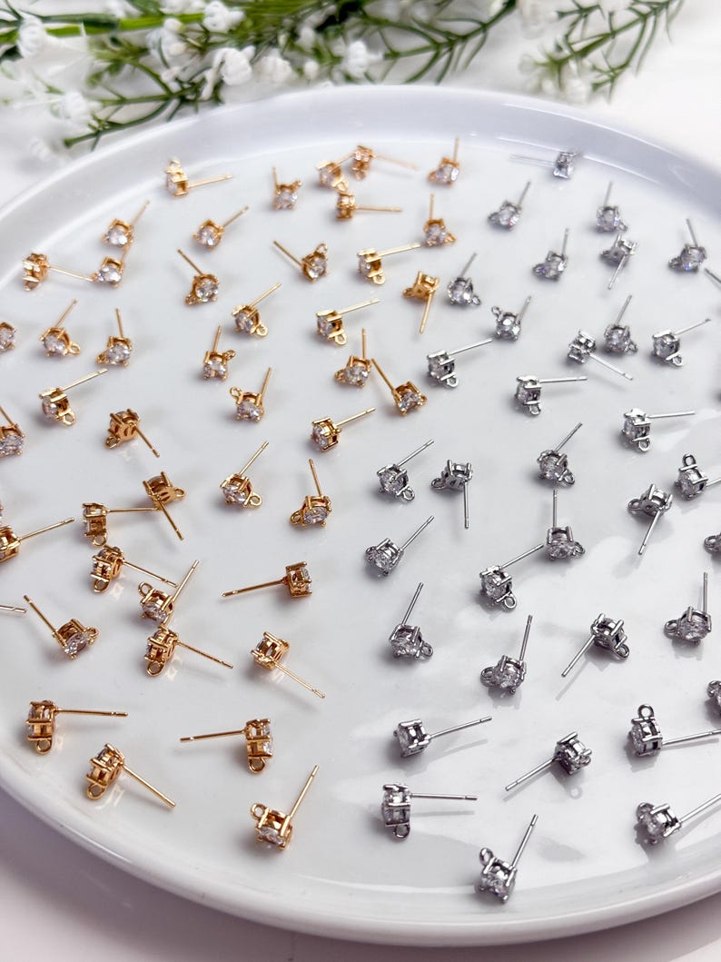May include: A collection of gold-toned and silver-toned stud earrings, each featuring a single clear round gemstone set in a four-prong metal basket. Each earring includes a small loop at the base of the setting for attaching additional charms.