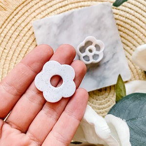 Op de afbeelding: Witte, gevlekte koekjesvormer in de vorm van een bloem met een gat in het midden. De vormer wordt in een hand gehouden.