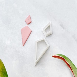 Può includere: Un set di quattro tagliabiscotti in plastica bianca a forma di diamanti geometrici. I tagliabiscotti sono su una superficie di marmo bianco. Uno dei tagliabiscotti è riempito di pasta di zucchero rosa.