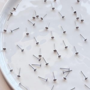 May include: A selection of silver earring posts with flat, circular backs, arranged on a white, oval ceramic plate. The posts are evenly distributed, forming a pattern against the bright background. The earrings are small and delicate.