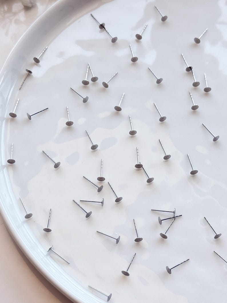 May include: A selection of silver earring posts with flat, circular backs, arranged on a white, oval ceramic plate. The posts are evenly distributed, forming a pattern against the bright background. The earrings are small and delicate.