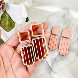 以下が含まれることがあります： 窓の形をしたプラスチック製のクッキーカッター5個セット。カッターはベージュで、滑らかな仕上がりです。正方形のカッターが2つ、縦線が引かれた長方形のカッターが2つ、横線が引かれた長方形のカッターが1つあります。カッターは手と大理石の表面の上に置かれています。ピンクでテクスチャのある正方形のフォンダンが4つもあります。