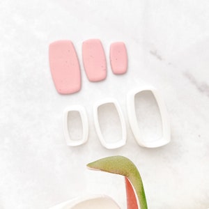 May include: Set of five white plastic cookie cutters in different sizes, shaped like rectangles. Three pink rectangles of fondant are also shown.