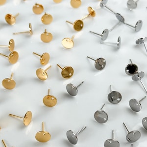 Puede incluir: Una colección de pendientes de botón de metal en tonos dorados y plateados, cada uno con un disco circular plano y una pequeña anilla en la base para su fijación. Los pendientes están dispuestos de forma dispersa, mostrando ambos acabados.