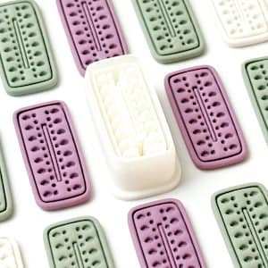 Puede incluir: Un surtido de moldes de jabón rectangulares decorativos en varios colores, incluyendo verde, morado y blanco. Los moldes tienen un patrón punteado y una ranura central. Un molde blanco está abierto, revelando el interior del jabón.