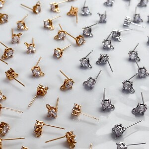 May include: A collection of stud earrings featuring clear, round-cut stones set in gold-tone and silver-tone metal. Each earring includes a post back and a small loop at the base of the setting, arranged in a scattered pattern across the surface.
