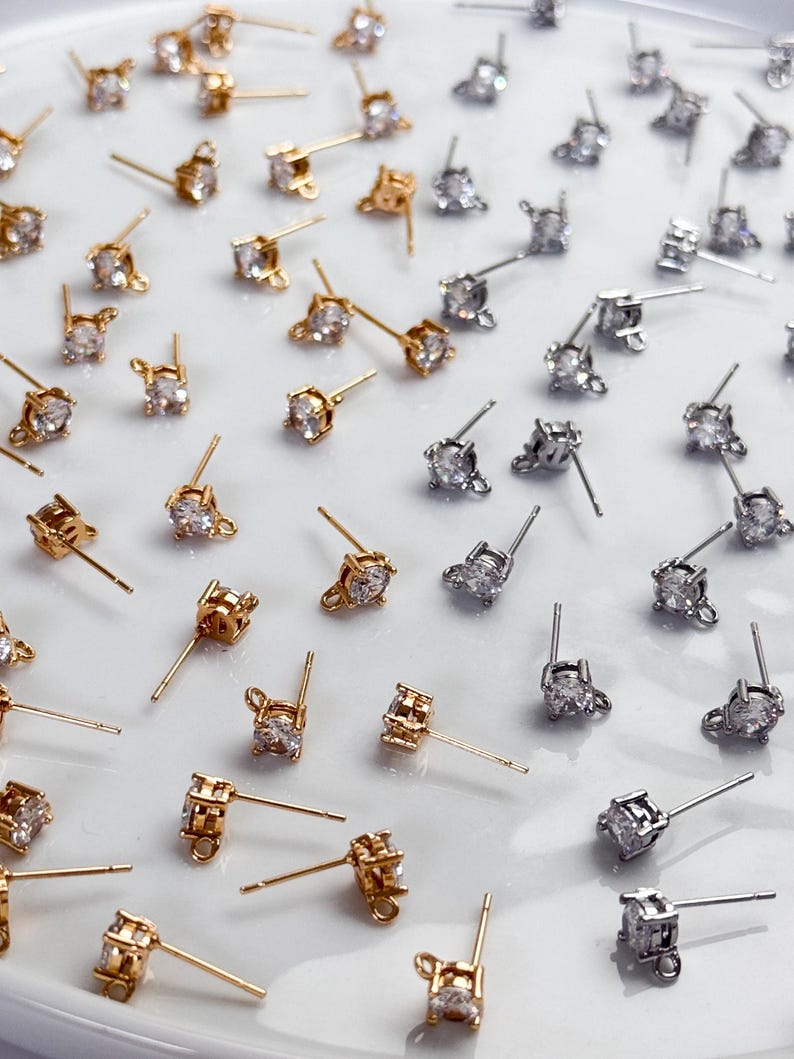 May include: A collection of stud earrings featuring clear, round-cut stones set in gold-tone and silver-tone metal. Each earring includes a post back and a small loop at the base of the setting, arranged in a scattered pattern across the surface.