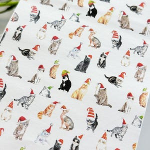 Puede incluir: Tela blanca con un patrón repetido de gatos con gorros de Papá Noel. Los gatos están en varias poses y colores, incluyendo negro, blanco, gris, marrón y naranja.