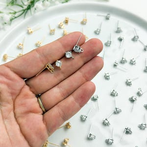 May include: A collection of stud earring findings featuring a four-prong setting with a clear round stone and a small loop at the base. The findings are available in both gold and silver tones, with many pieces scattered across a white surface.