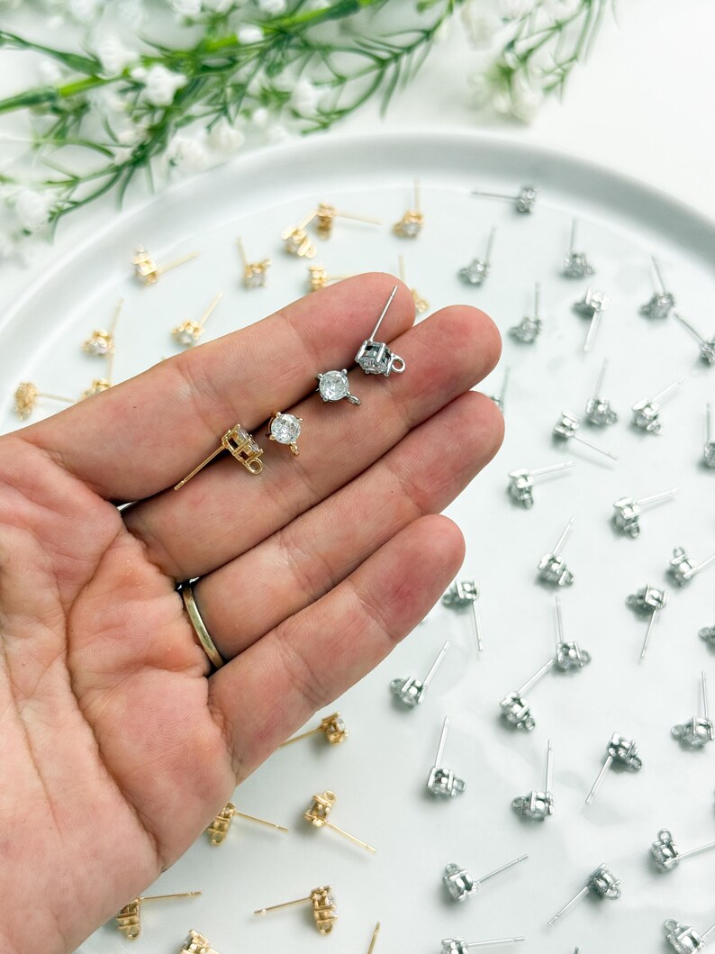 May include: A collection of stud earring findings featuring a four-prong setting with a clear round stone and a small loop at the base. The findings are available in both gold and silver tones, with many pieces scattered across a white surface.