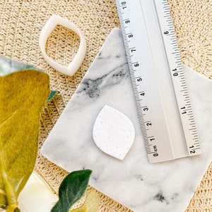 Puede incluir: Una esponja de maquillaje blanca en forma de hoja con textura moteada, un anillo de plástico blanco y una regla plateada con marcas en centímetros y pulgadas.