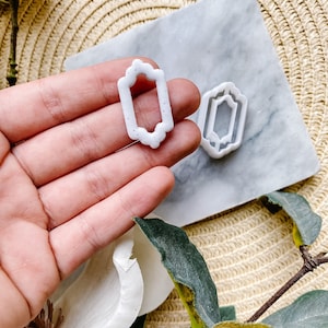 May include: Two white plastic cookie cutters in the shape of a rectangle with ornate edges. The cutters are on a marble surface with a woven mat in the background.