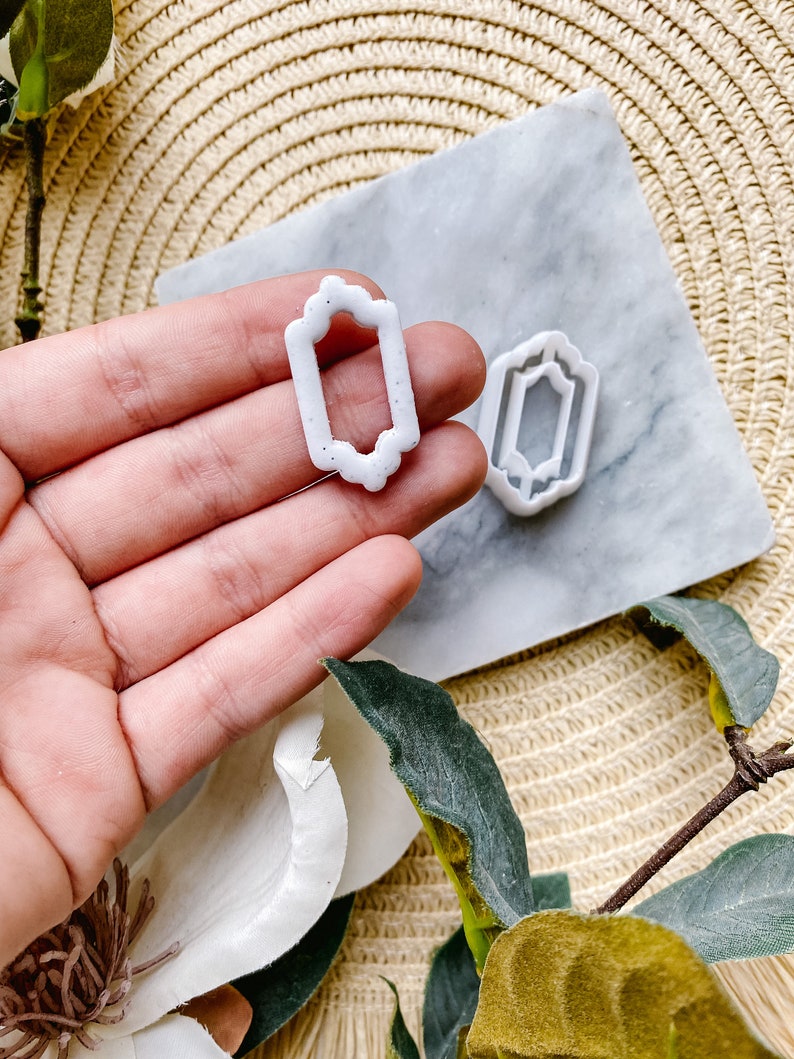 May include: Two white plastic cookie cutters in the shape of a rectangle with ornate edges. The cutters are on a marble surface with a woven mat in the background.