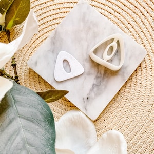 Puede incluir: Dos cortadores de galletas de plástico blanco en forma de triángulo sobre una superficie de mármol blanco. Los cortadores de galletas tienen forma de triángulo con la parte superior redondeada y un agujero en el centro.