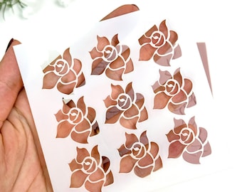 May include: A sheet of stencils featuring nine rose designs. The stencils are cut out of a translucent white material, revealing the rose pattern in a warm, reddish-brown hue. The stencils are held in a hand against a white background.