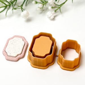 Op de afbeelding: Drie koekjessnijders in een decoratieve vorm. Eén is roze met een gespikkeld wit interieur, en de andere twee zijn tinten oranje. De uitstekers zijn gerangschikt op een wit oppervlak met witte bloemen op de achtergrond.