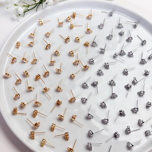 May include: A collection of small stud earring findings arranged on a white plate. The set includes gold-toned and silver-toned metal posts, each featuring a single clear cubic zirconia stone and a small loop at the base for attaching charms.