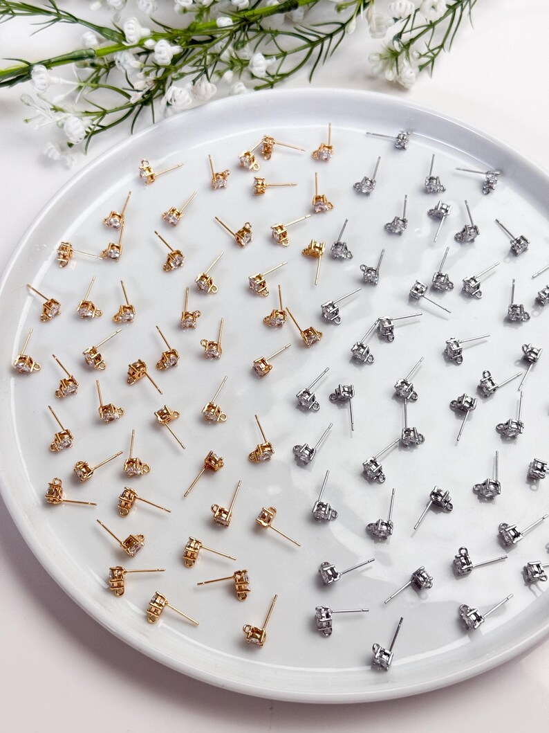 May include: A collection of small stud earring findings arranged on a white plate. The set includes gold-toned and silver-toned metal posts, each featuring a single clear cubic zirconia stone and a small loop at the base for attaching charms.