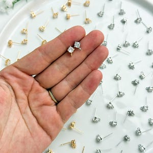 May include: A collection of small stud earrings featuring clear round stones set in metal prongs. The earrings are available in gold and silver tones, each with a thin post and a small loop at the base for attaching additional jewellery components.