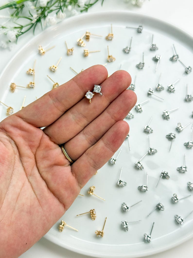 May include: A collection of small stud earrings featuring clear round stones set in metal prongs. The earrings are available in gold and silver tones, each with a thin post and a small loop at the base for attaching additional jewellery components.