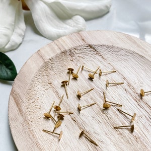 May include: A close-up of a wooden plate with a pile of gold earring backs. The backs are small and round with a small pin attached to the back.
