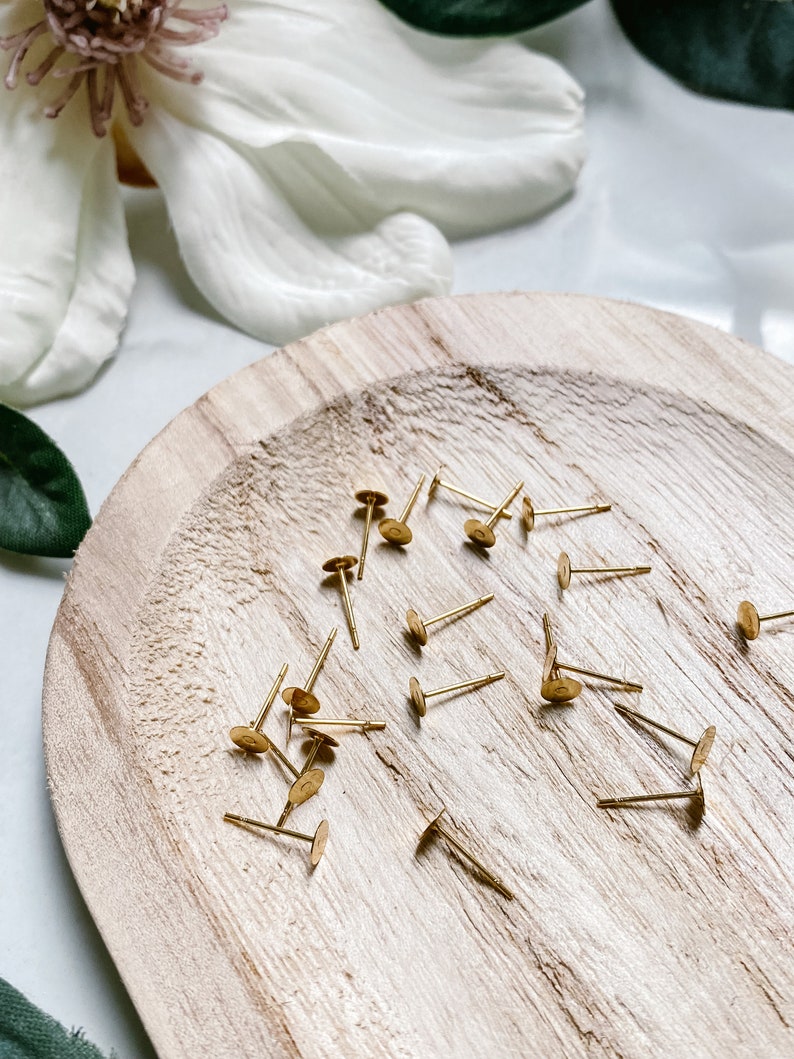 May include: A close-up of a wooden plate with a pile of gold earring backs. The backs are small and round with a small pin attached to the back.