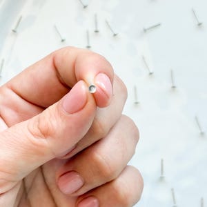 May include: A tiny, silver stud earring with a clear, round gem is held between fingers. The background shows more silver studs on a white surface, indicating a jewellery or craft theme.