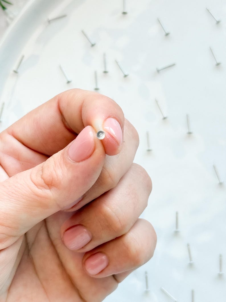 May include: A tiny, silver stud earring with a clear, round gem is held between fingers. The background shows more silver studs on a white surface, indicating a jewellery or craft theme.