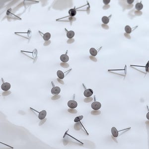 May include: A collection of silver earring posts with flat, round pads and thin posts, scattered on a white surface. These earring posts are for jewellery making and crafting.