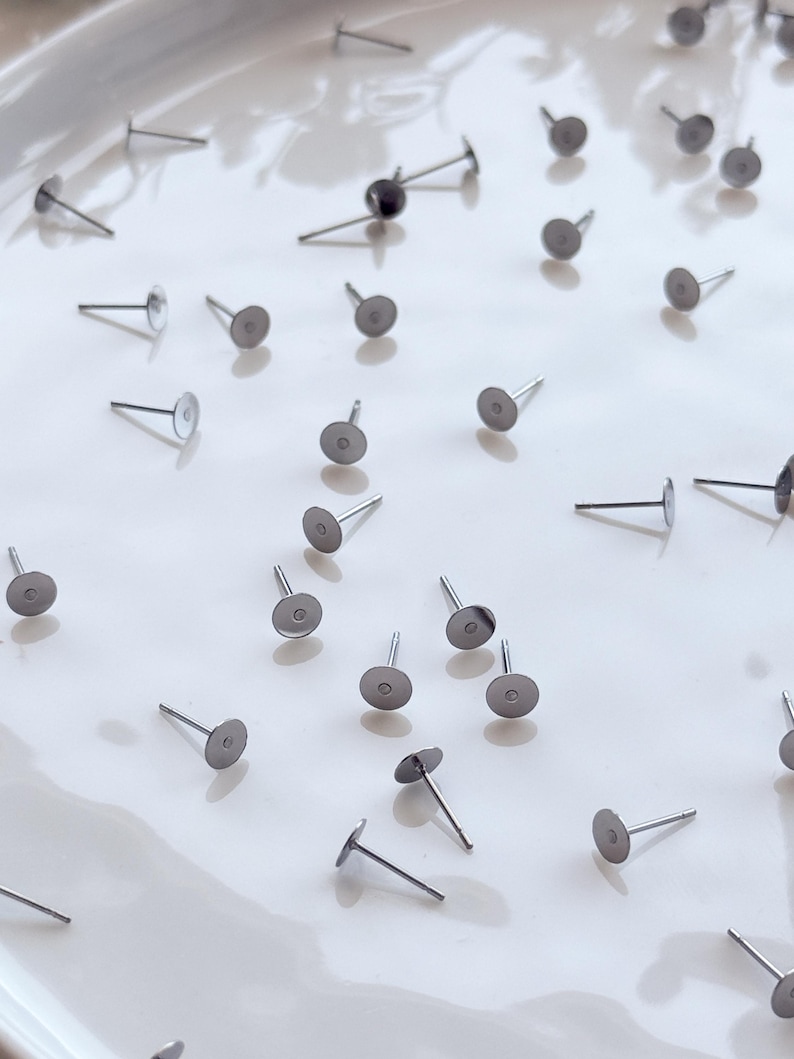May include: A collection of silver earring posts with flat, round pads and thin posts, scattered on a white surface. These earring posts are for jewellery making and crafting.