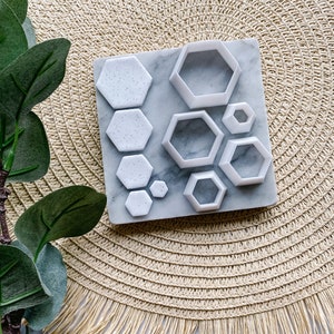 May include: A selection of white, hexagon-shaped cookie cutters and matching speckled clay tiles on a marble surface. The cutters are of varying sizes, and the tiles have a speckled pattern. The items are on a beige woven placemat.
