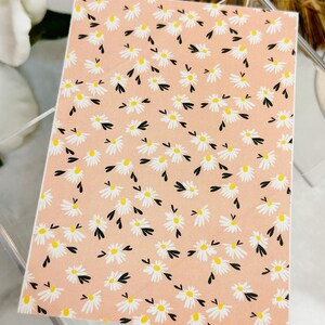 May include: A pink background with a repeating pattern of white daisies with yellow centres and black stems. The daisies are scattered randomly across the background.