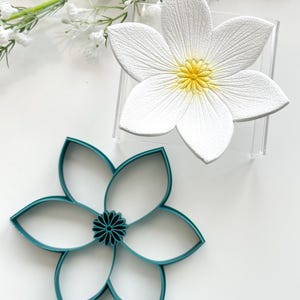 Peut inclure: Une fleur en céramique blanche avec un centre jaune et un emporte-pièce en plastique turquoise en forme de fleur. L'emporte-pièce a six pétales.